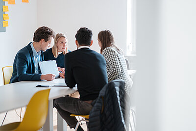 Personen in einem Meeting am Tisch, fokussiert und diskussionierend, mit Notizzetteln an der Wand im Hintergrund und warmem Tageslicht im Raum.