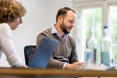 Zwei Personen in einem Besprechungsraum konzentriert an einem Laptop arbeitend, im Hintergrund eine helle Fensterfront und Glasflaschen.