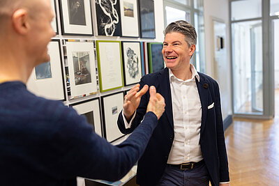 Zwei Männer in einem Büro lächeln und unterhalten sich vor einer Wand mit gerahmten Kunstwerken bei Undconsorten.